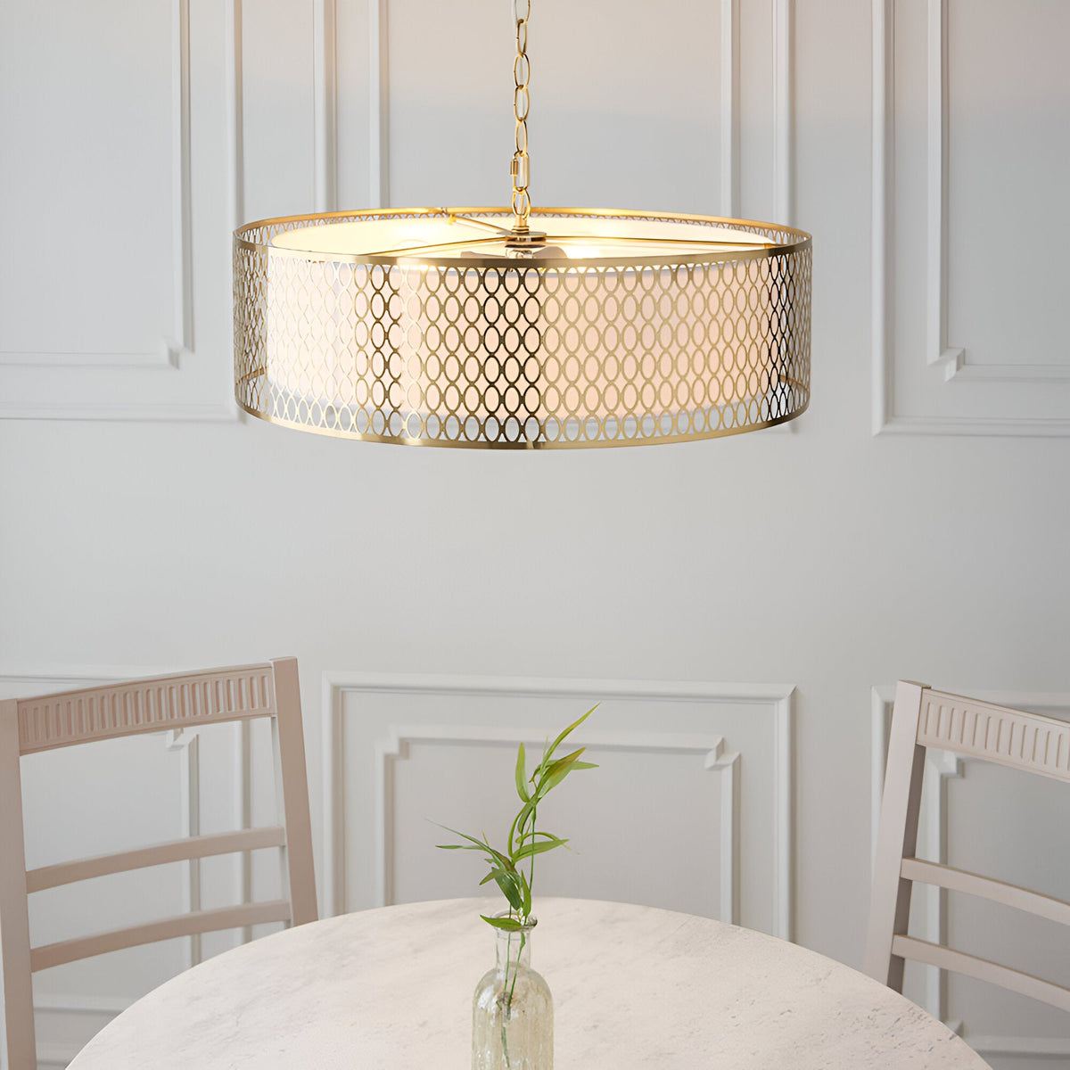 Rosaline 3 Light Pendant in gold with circular fretwork outer and white fabric inner shade, hanging above a dining table in a light-panelled room.