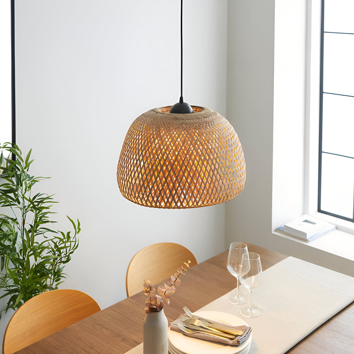 Aldstead Oval Pendant in natural woven rattan with matt black fittings, hung above a wooden dining table in a bright room setting.