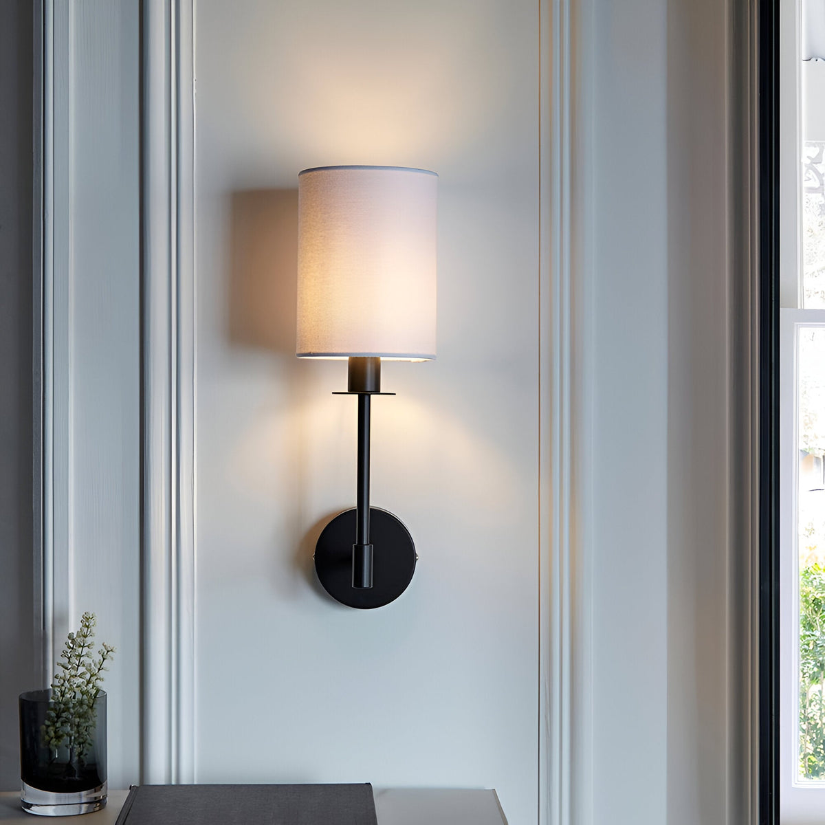 Endon Cliffhurst wall light with a slim matt black upright and vintage white cylinder shade, mounted beside a window above a console with a vase.