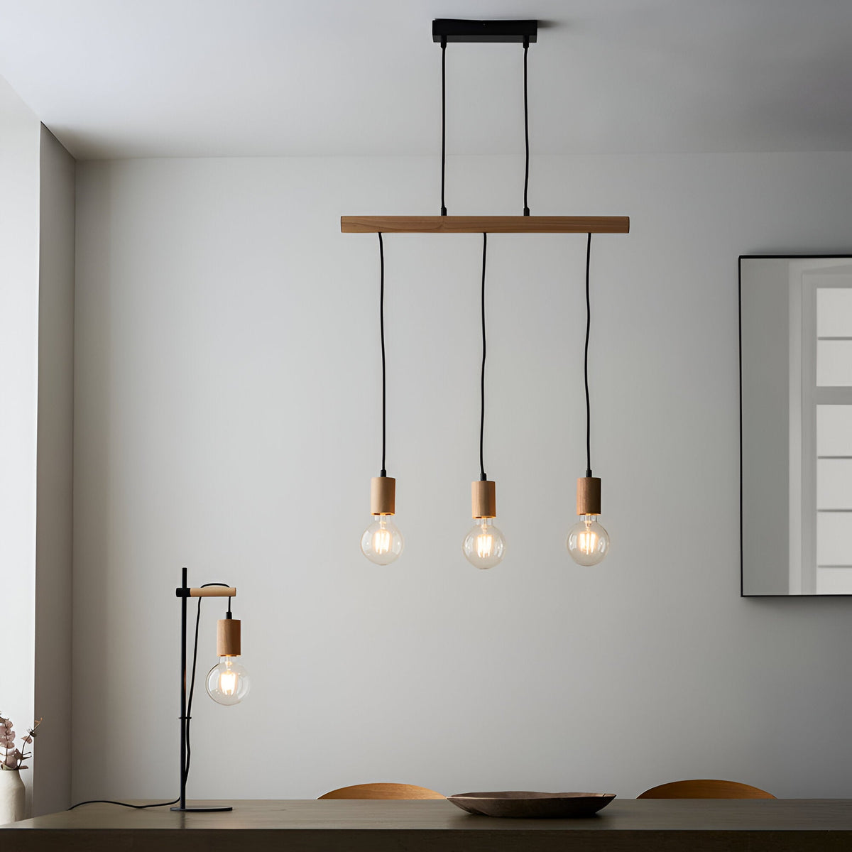 Cranbury 3-Light Linear Pendant in black and natural wood with staggered exposed filament bulbs, shown above a dining table beside a matching table lamp.