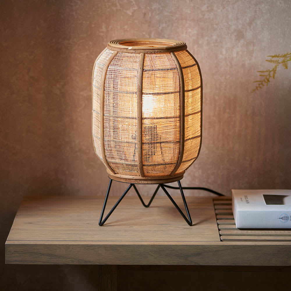 Endon Daneshaw table lamp with woven natural linen and bamboo shade on matt black metal legs, lit on a wooden console against a warm earthy wall.