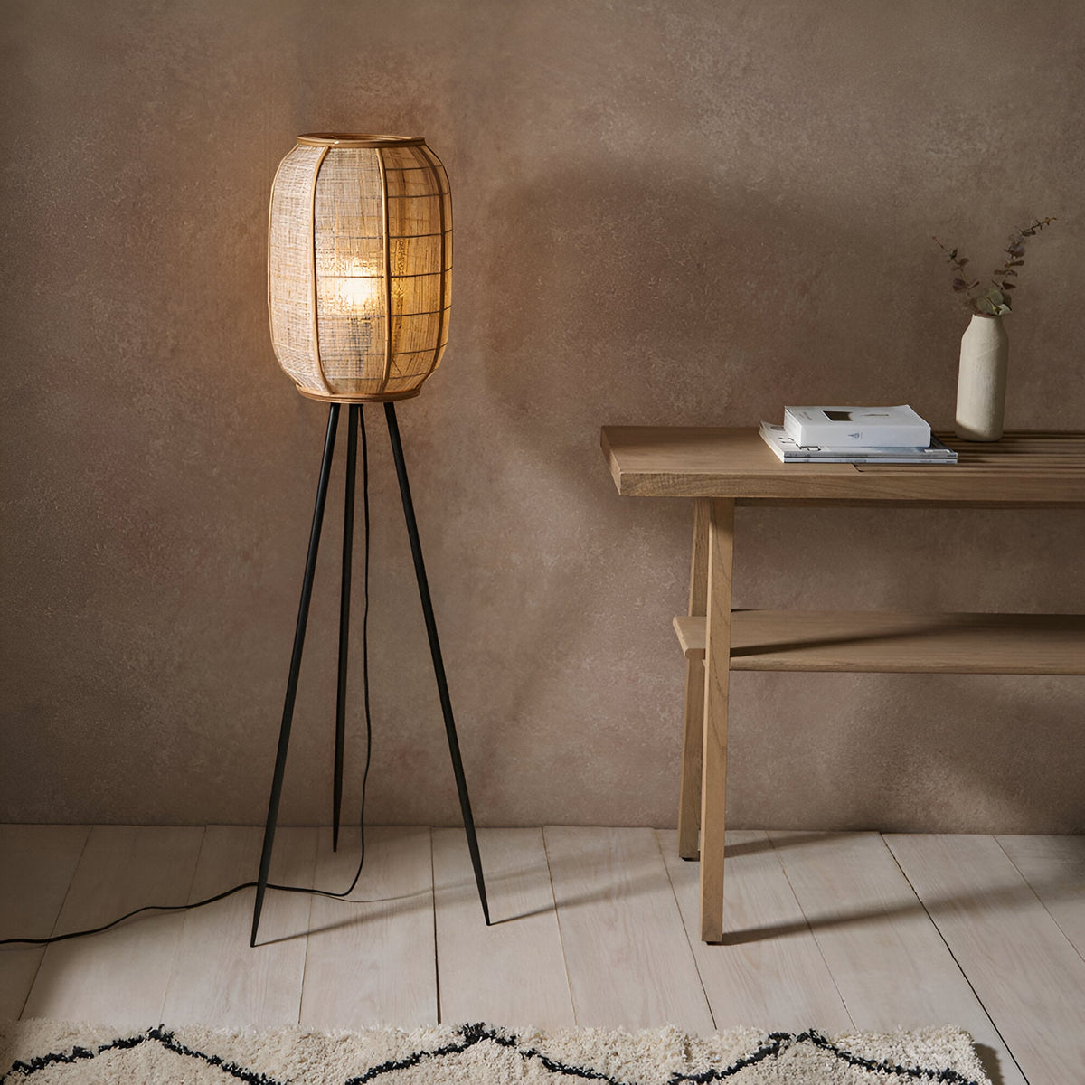Daneshaw Floor Lamp with woven natural linen shade on a bamboo frame and black tripod base, glowing beside a wooden console table.