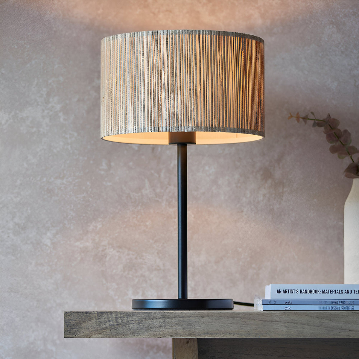 Salcombe Table Lamp with woven seagrass drum shade on a matt black metal base and stem, shown lit on a wooden console table.