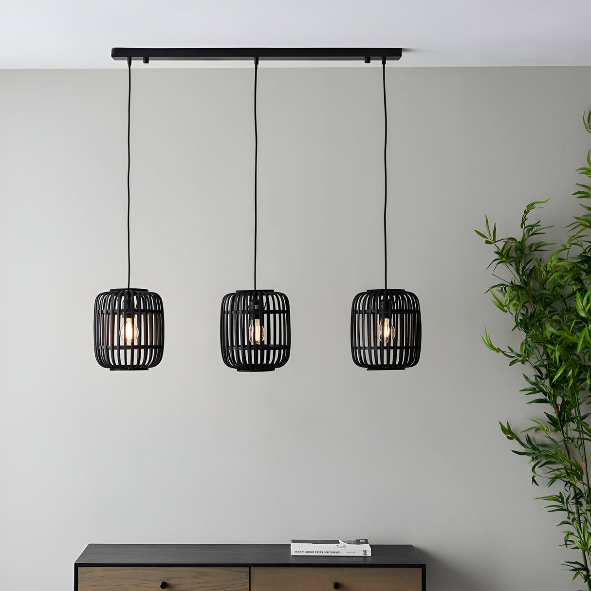 Endon Bracken 3 light linear pendant with black bamboo cage shades and matt black bar, hung above a dark sideboard beside a bamboo plant.