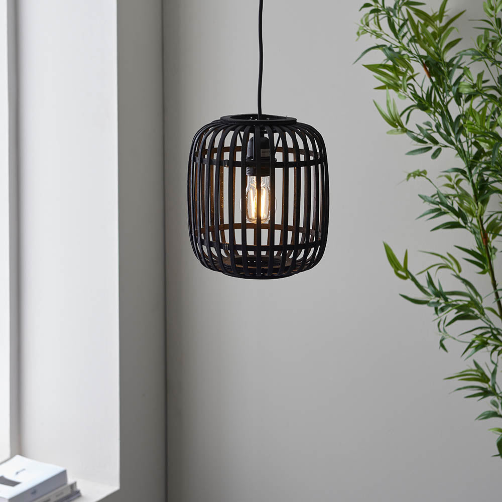 Bracken Pendant Light in black bamboo cage design with visible E27 filament bulb, hanging from black flex in a room with a green plant.