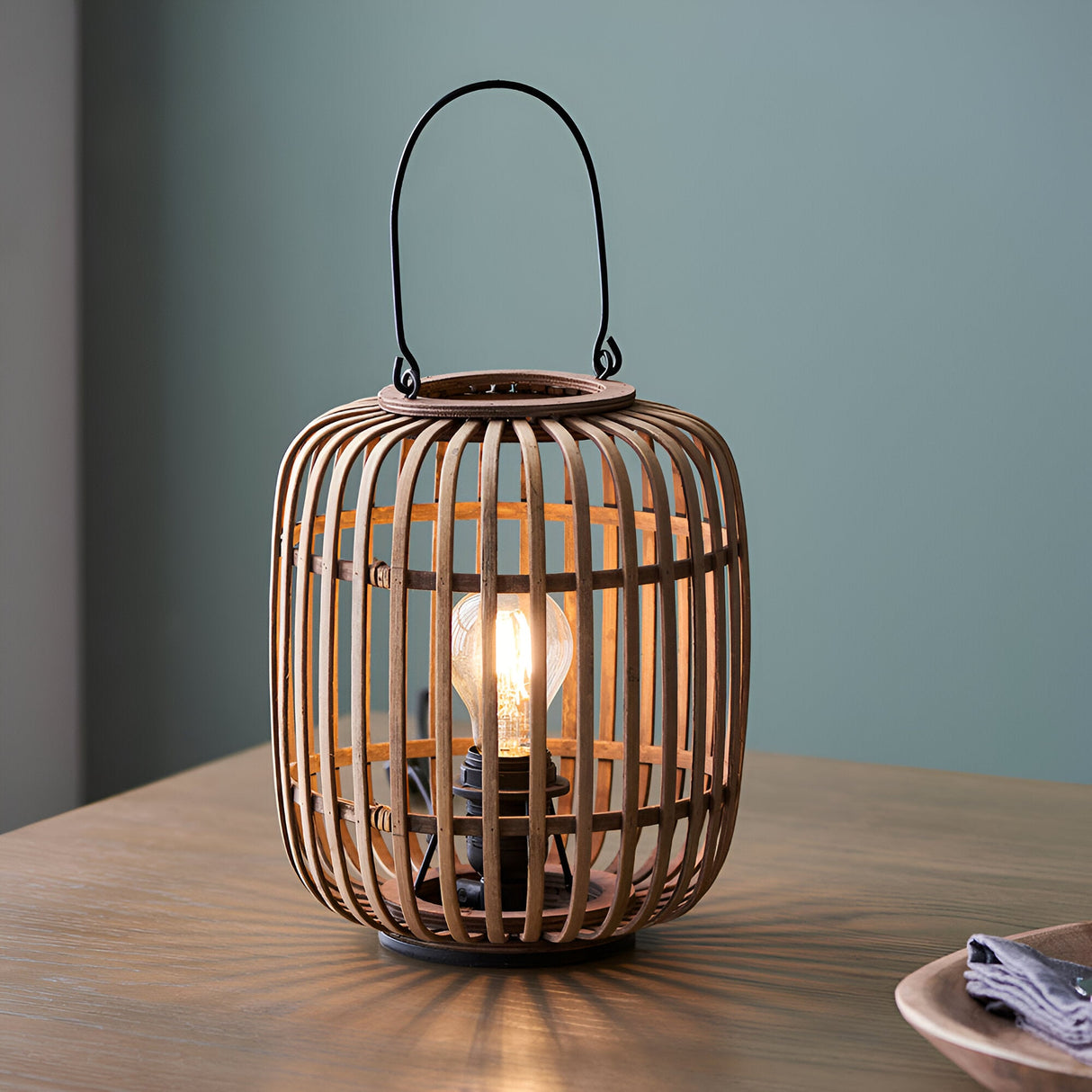 Endon Bracken table lamp with an open bamboo cage, matt black metal handle and exposed E27 filament bulb, shown lit on a wooden table.