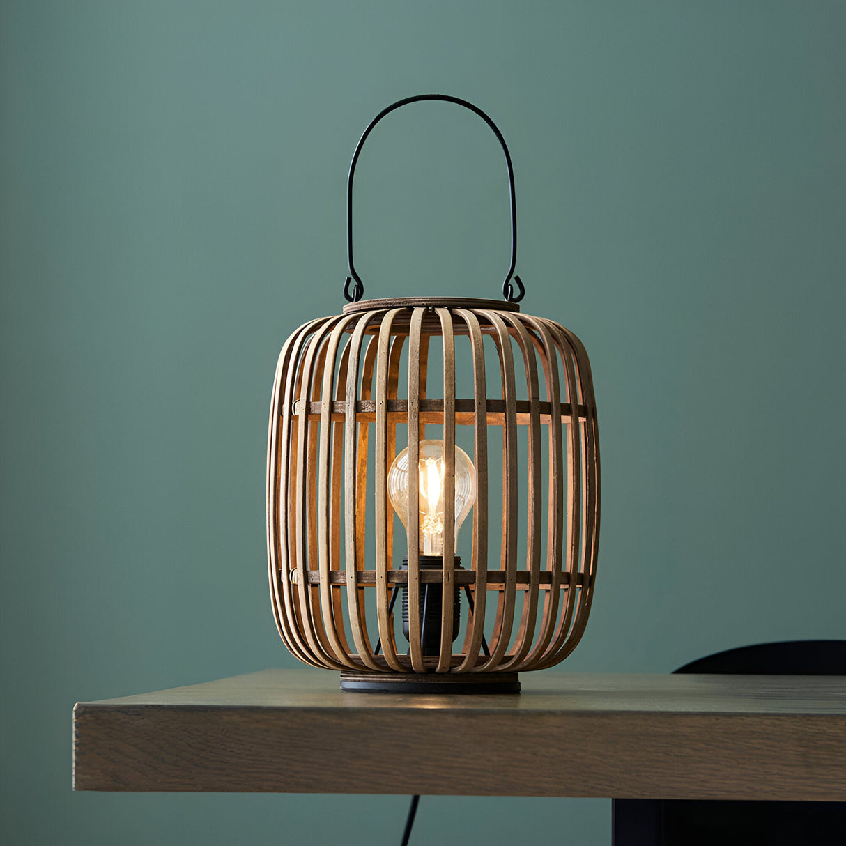 Endon Bracken table lamp with natural bamboo cage, matt black metal handle and filament E27 bulb, on a wooden table against a teal wall.