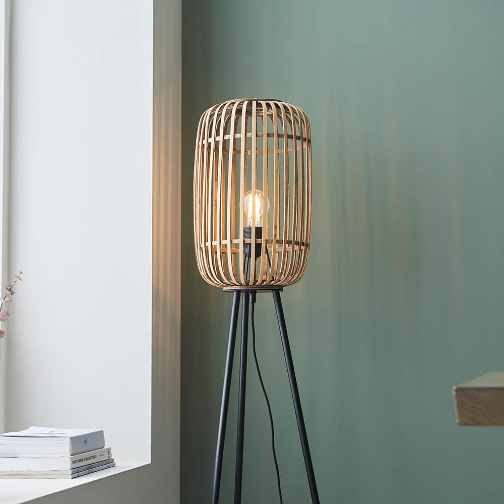 Bracken Floor Lamp with natural bamboo cage shade and black tripod base, shown lit in a living room against a green wall.