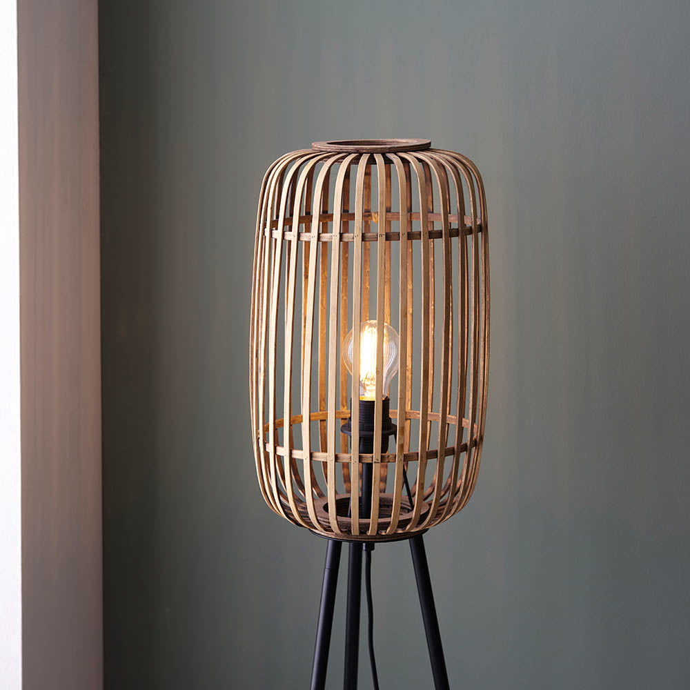 Bracken Floor Lamp in black and natural, showing a cylindrical bamboo cage shade with visible bulb on a black tripod base, set against a grey wall.