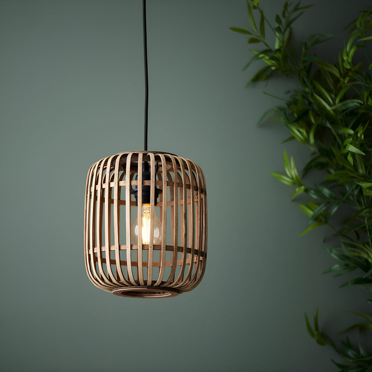 Endon Bracken pendant light with a natural bamboo slatted cage shade and matt black fitting, illuminated by an exposed filament bulb against a green wall.