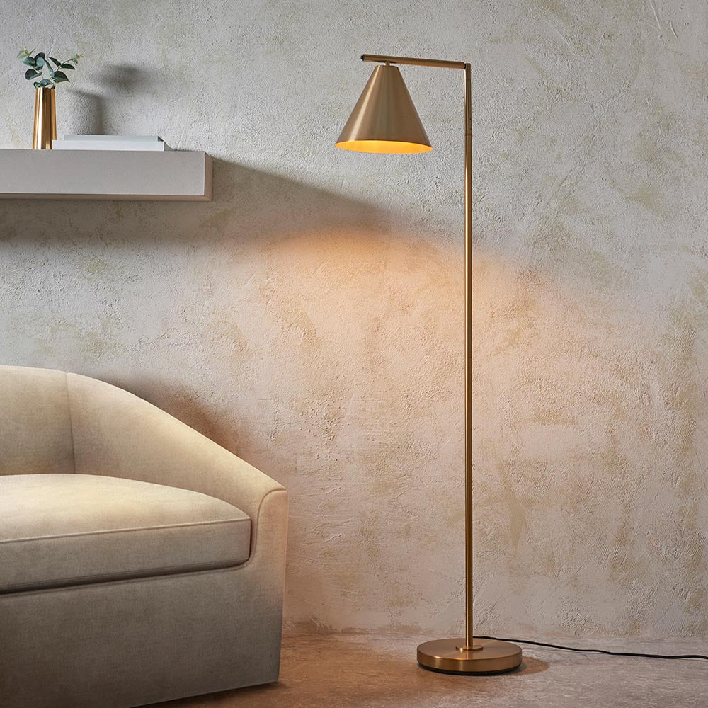Endon Ashworth brass floor lamp with adjustable arm and conical shade, illuminated beside a cream armchair and wall shelf in a living room.