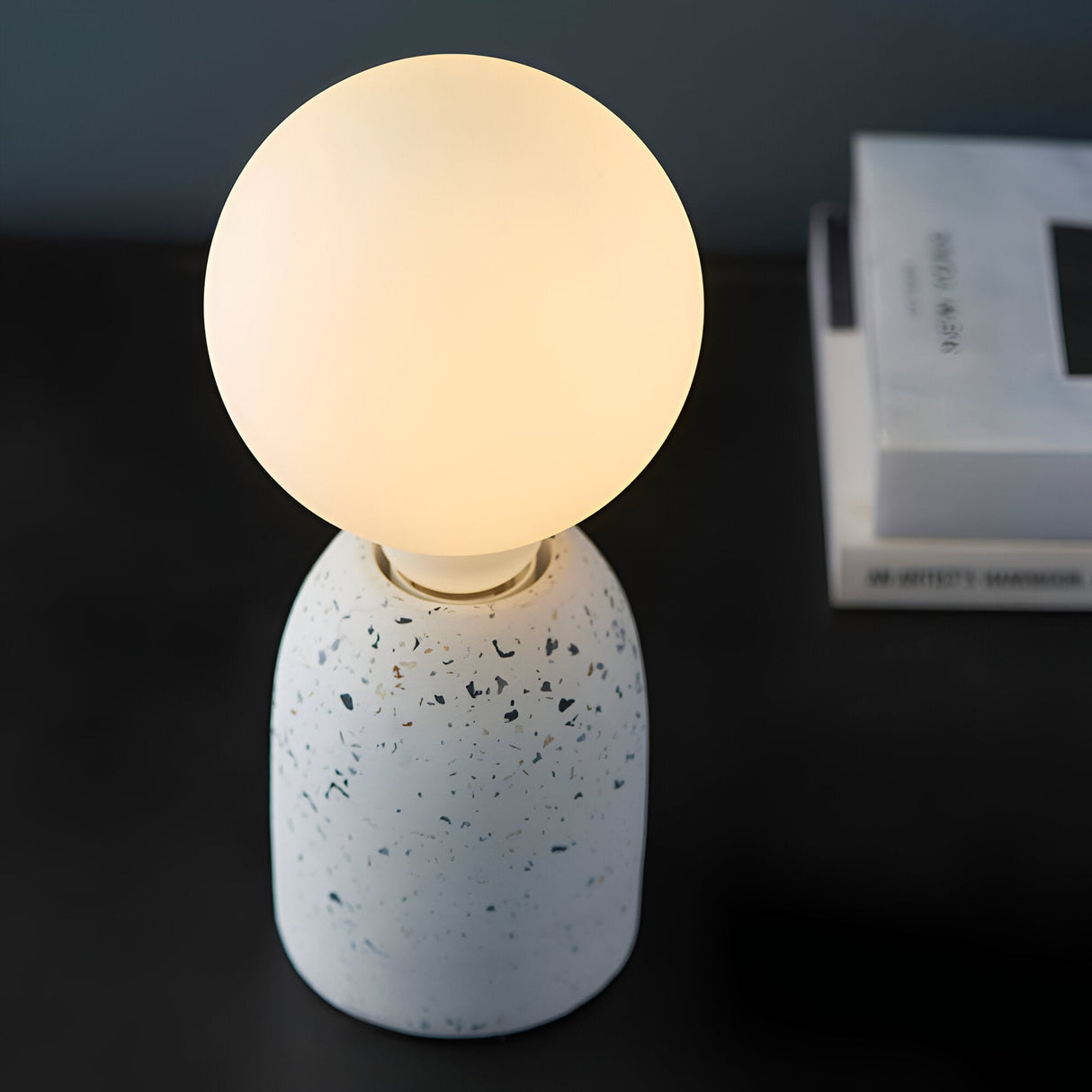 Endon 12W E27 LED filament globe bulb glowing warm white through opal glass on a terrazzo-style table lamp base beside stacked books.