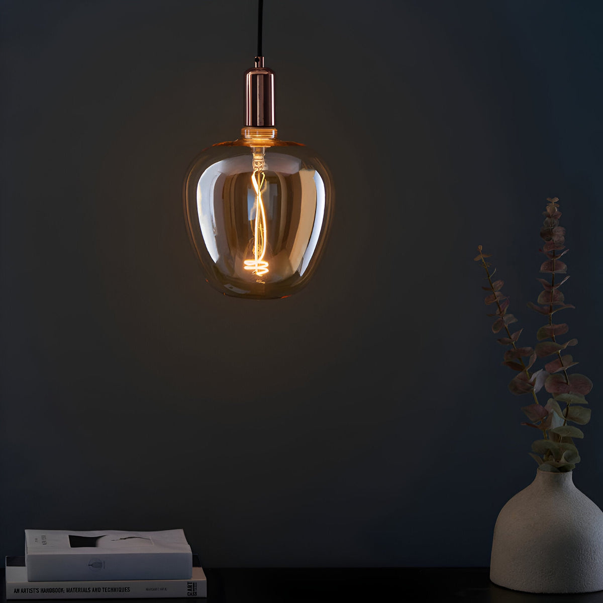 Endon 4W E27 LED filament swirl bulb with amber tinted glass and copper holder, hung on a black flex above a console with books and eucalyptus.