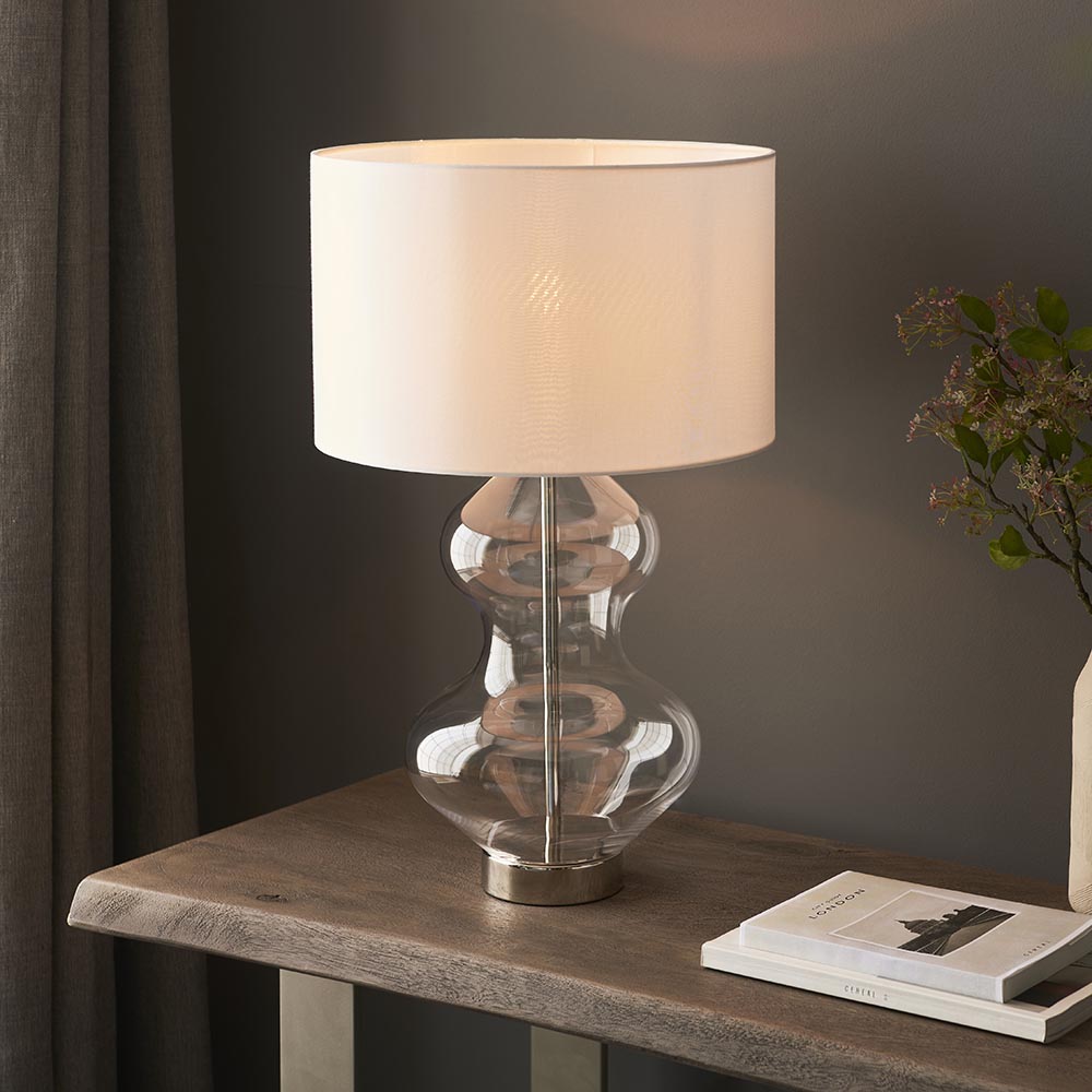 Endon Brockley table lamp with a clear glass hourglass base, polished nickel fitting and vintage white drum shade, on a wood console beside foliage.