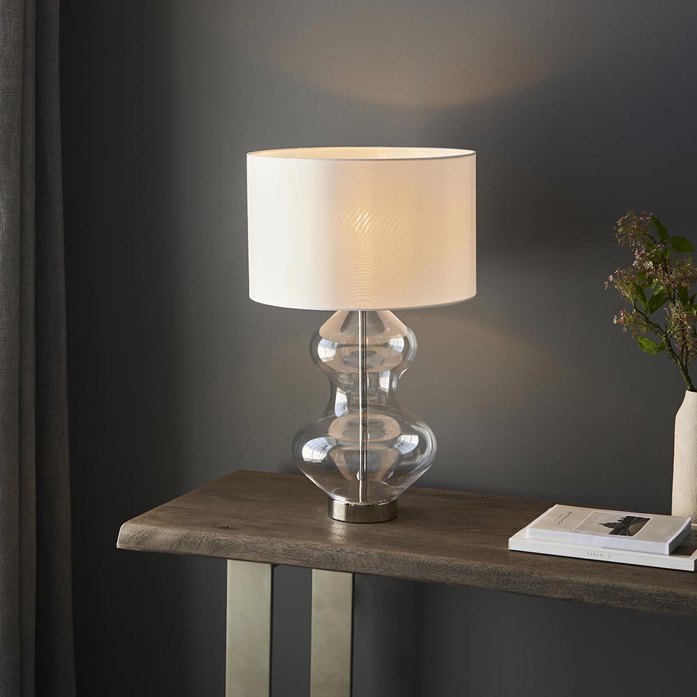 Endon Brockley table lamp with clear glass hourglass base, polished nickel fittings and white drum shade, on a wood console table against a grey wall.