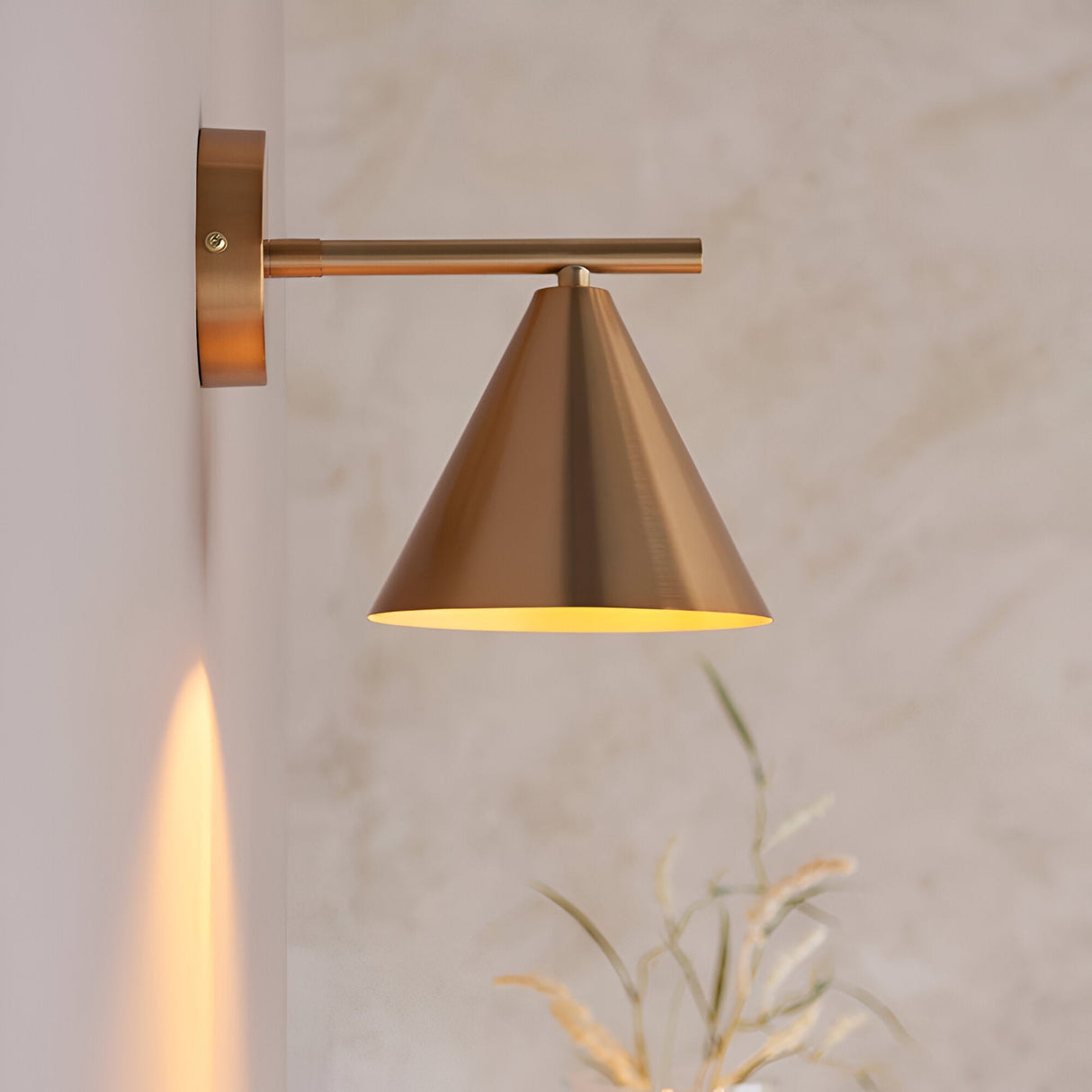 Endon Ashworth brass wall light with adjustable arm and conical shade, mounted on a light wall beside dried grasses, casting a warm glow.