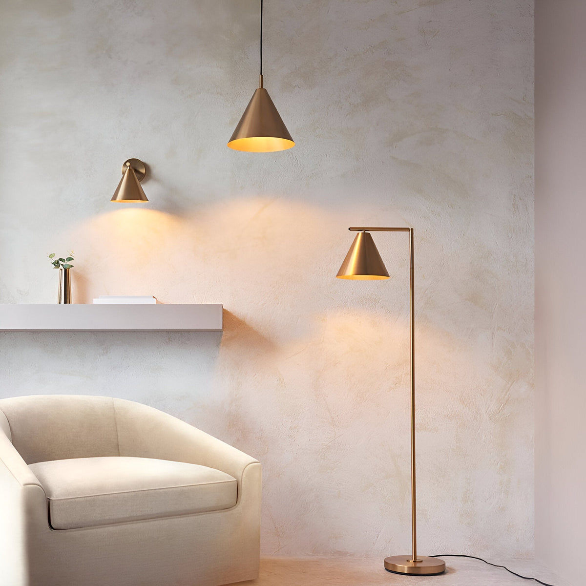 Endon Ashworth brass wall light with conical shade beside a matching pendant and floor lamp, cream armchair and shelf against a textured plaster wall.