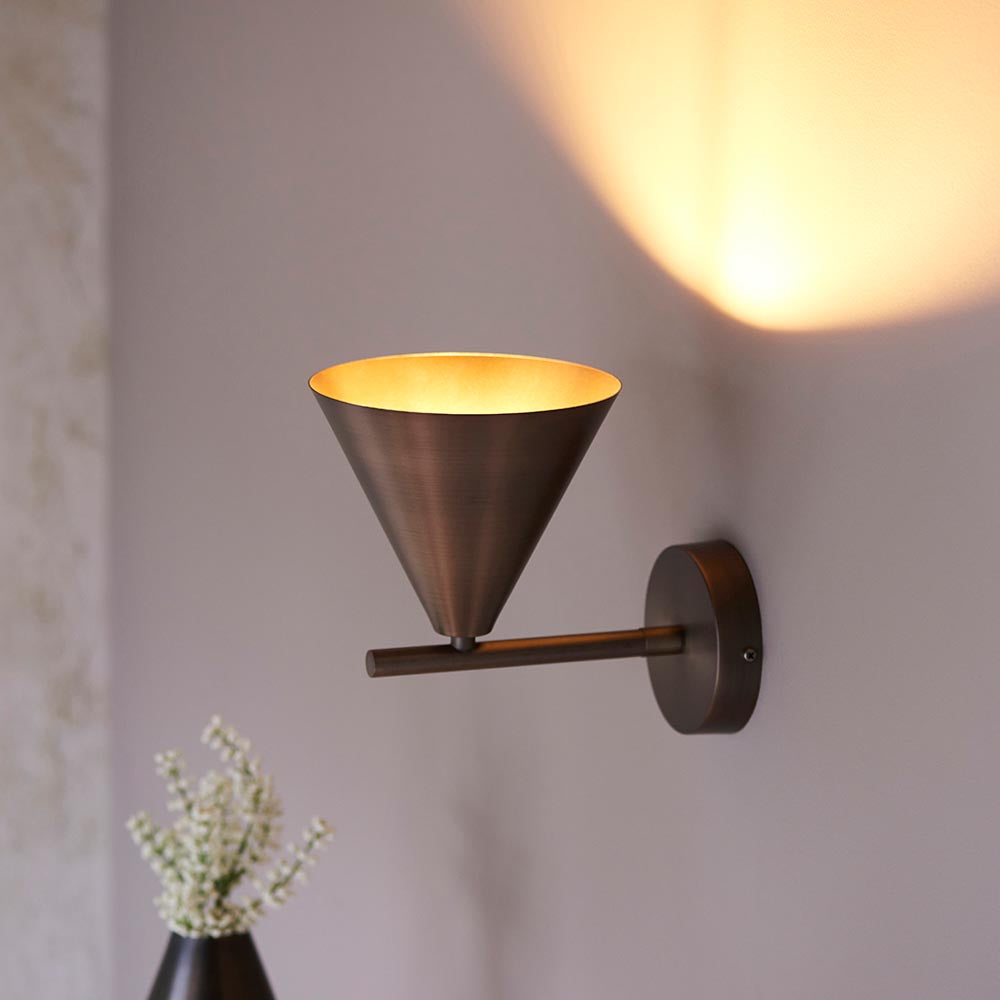 Endon Ashworth bronze wall light with a conical shade on an adjustable arm and round backplate, lit against a pale wall above white flowers.