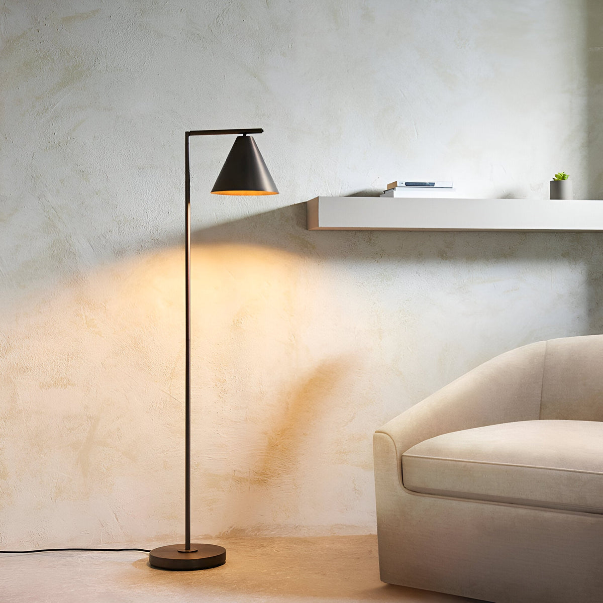 Ashworth Floor Lamp in bronze, single-light design with angled arm and cone shade, shown lit beside a sofa in a living room setting.