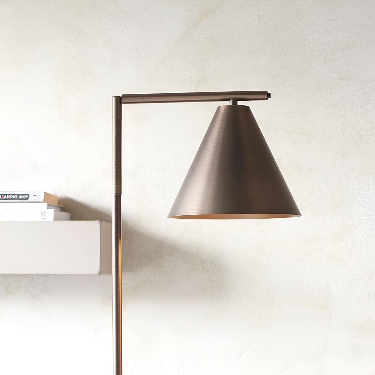 Ashworth Floor Lamp in bronze metal with adjustable arm and cone-shaped shade, shown beside a shelf of books against a light neutral wall.