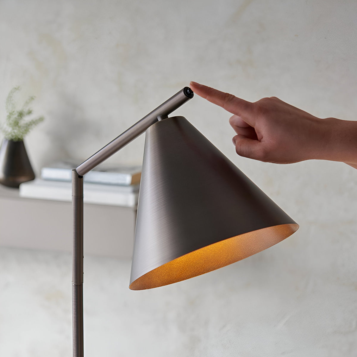 Ashworth Floor Lamp in bronze metal with a conical shade and adjustable angled arm, shown with a hand touching the push switch near a console table.
