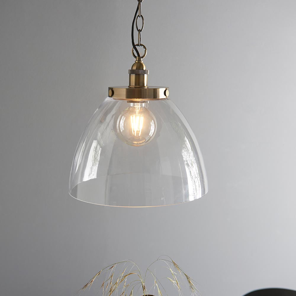Wentworth Pendant in brass with clear glass dome shade, knurled lampholder, and chain suspension, shown lit against a plain grey wall.