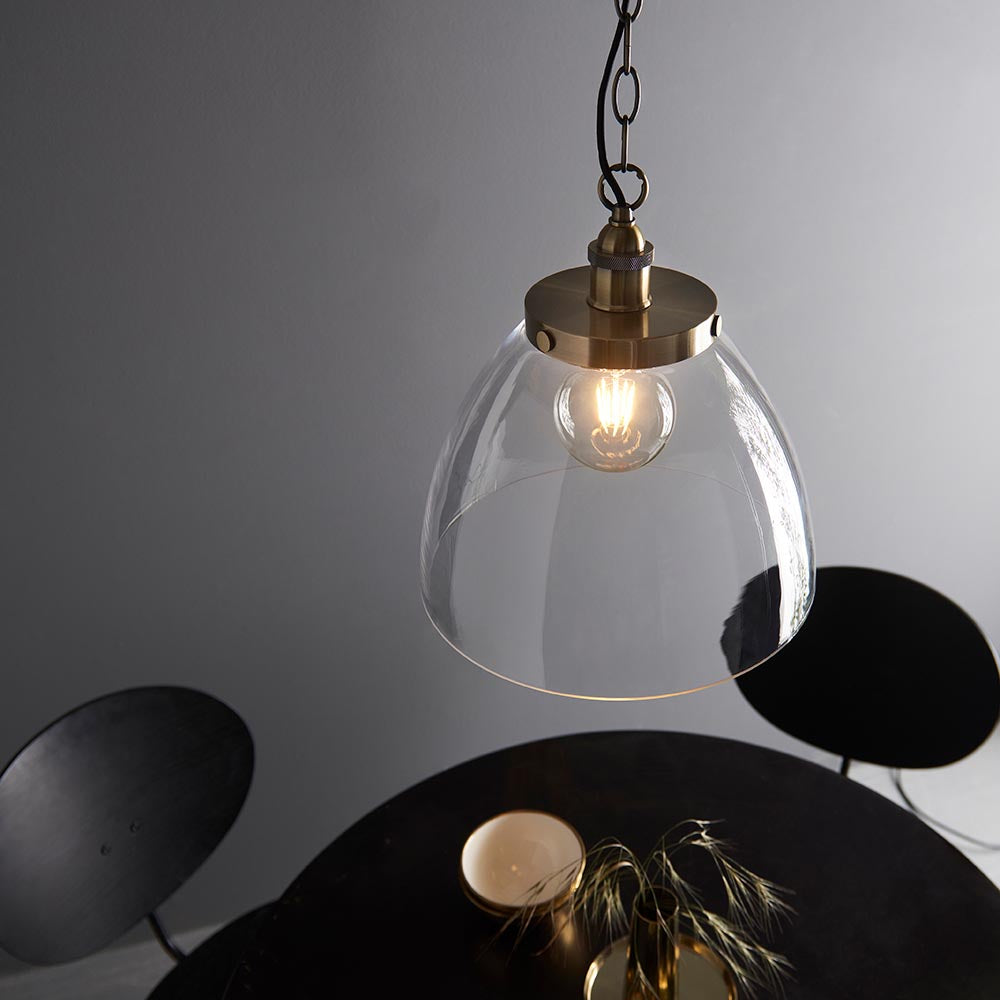 Wentworth Pendant in brass, industrial-style dome shade in clear glass with visible filament bulb, hung on a chain above a dark round table.