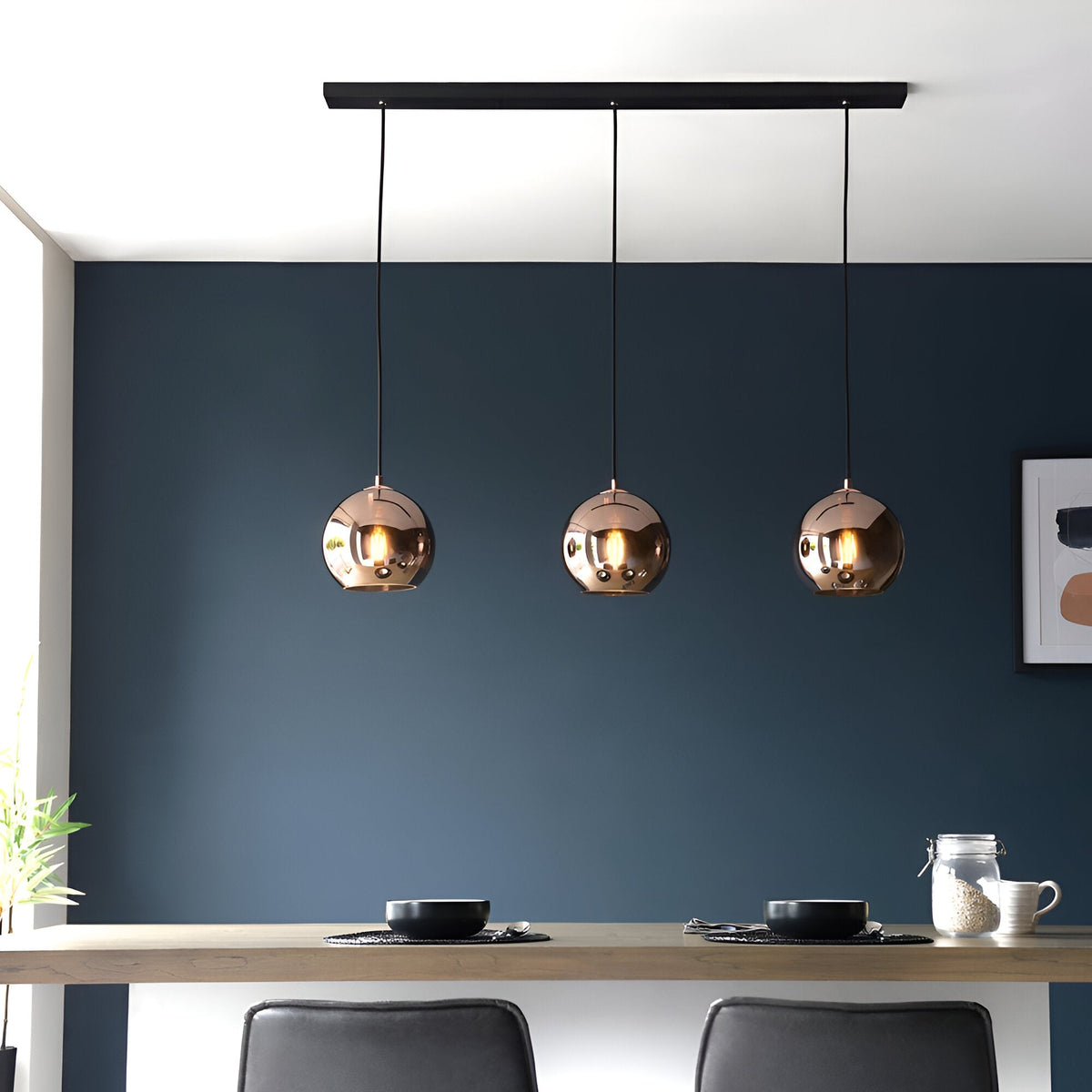 Endon Ashbourne 3 light linear pendant with copper mirrored glass globes on a matt black bar, hung above a dining table against a dark blue wall.