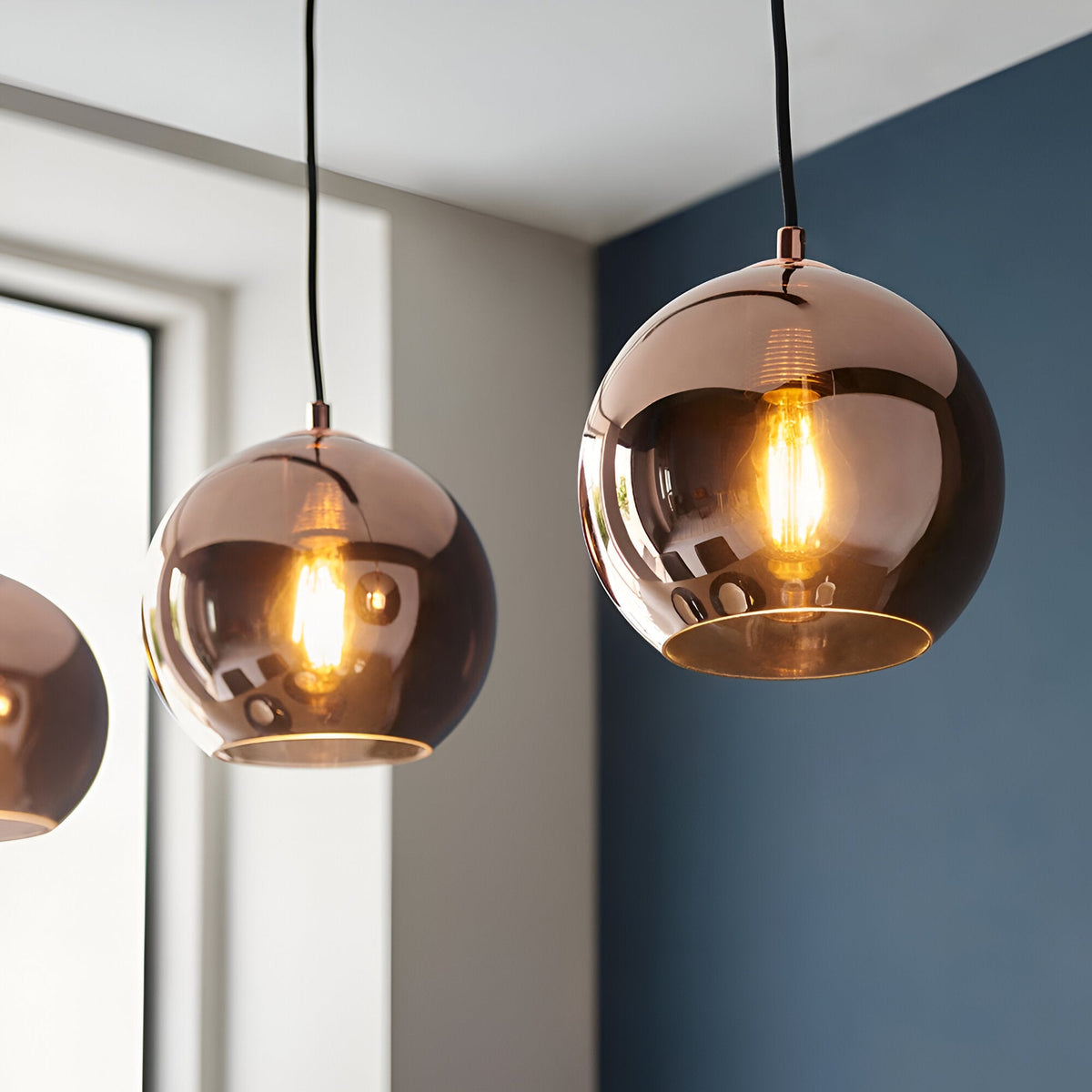Endon Ashbourne 3 light linear pendant with copper mirrored glass globe shades on black cables, shown lit in a room with blue and white walls.