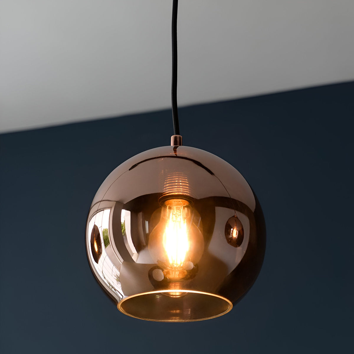 Endon Ashbourne pendant light with a single copper mirrored glass globe shade and exposed E27 filament bulb, hung from a black cable against a dark blue wall.