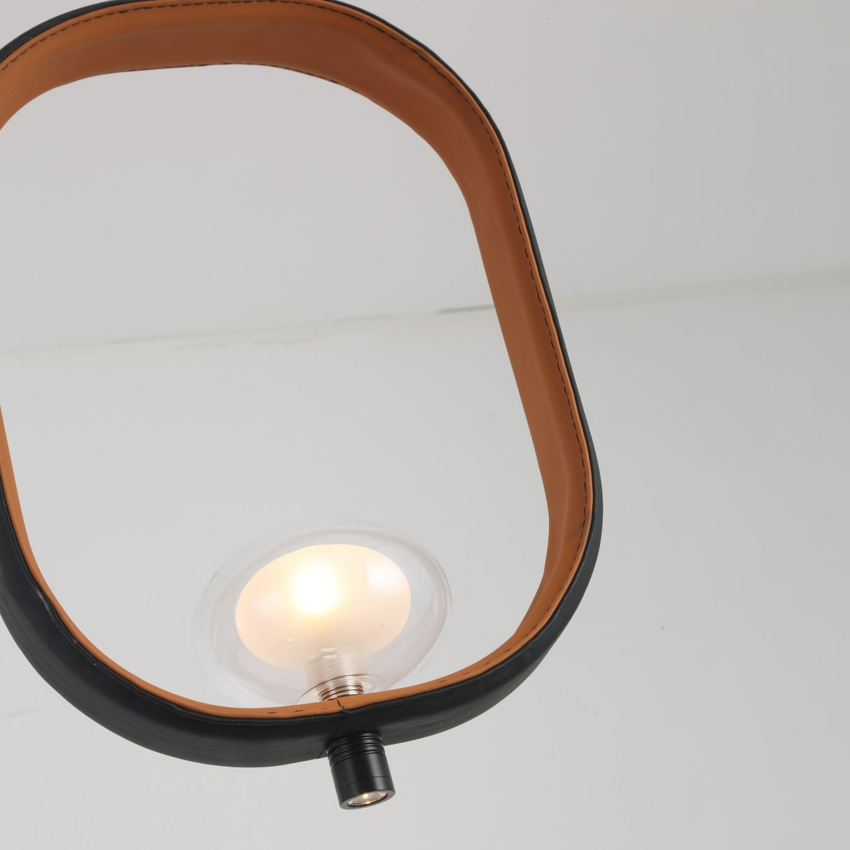 The Strappa Pendant Light - Grey/Black features a round, exposed bulb inside an oval or rectangular leather or faux leather frame, shown from below against a white ceiling—ideal as a modern LED pendant fixture.