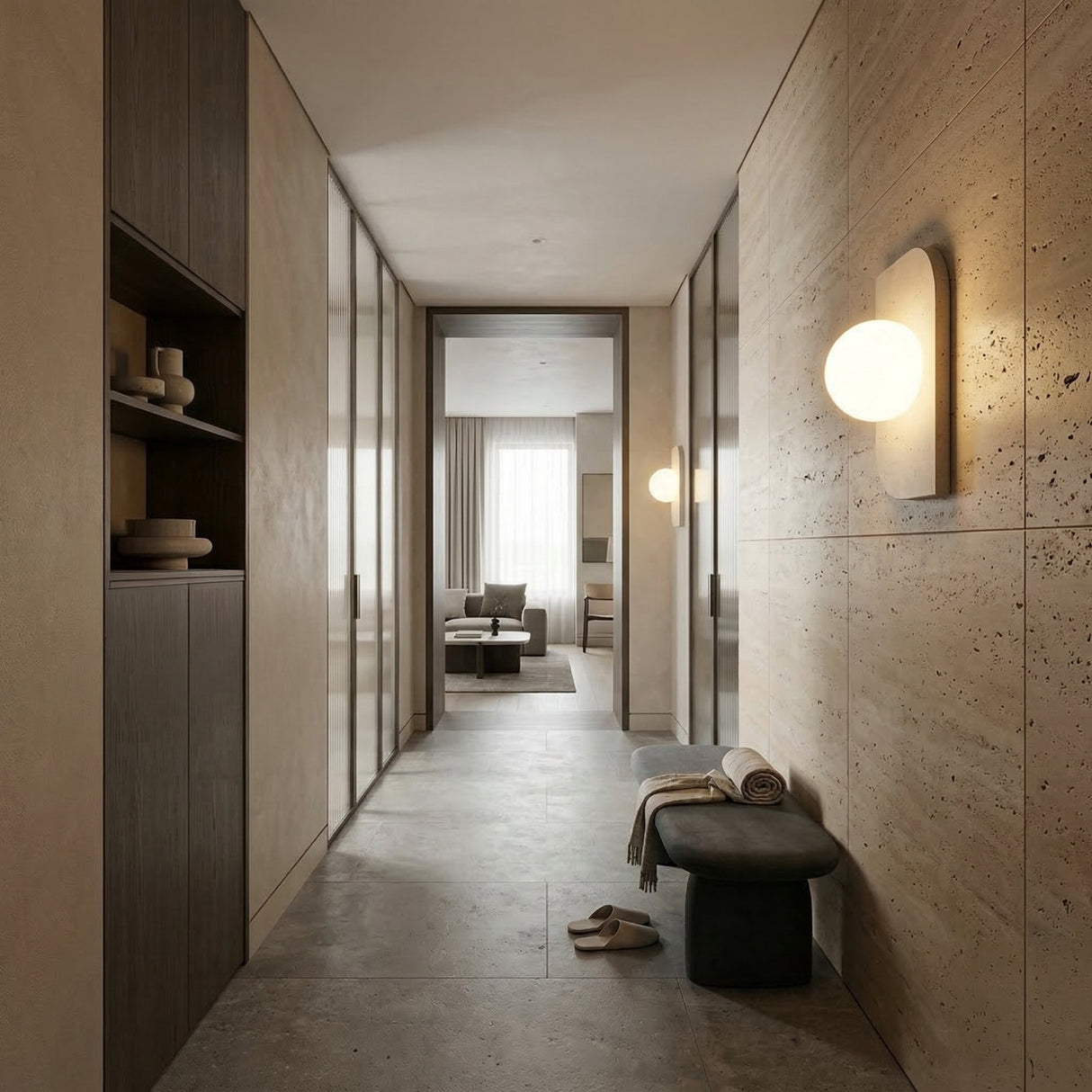 A modern, minimalist hallway with beige stone walls, built-in shelves, the Virella 1 Light Vertical Travertine Wall Light for ambient lighting, a bench with towels, and slippers leads to a bright living area with large windows and sheer curtains.