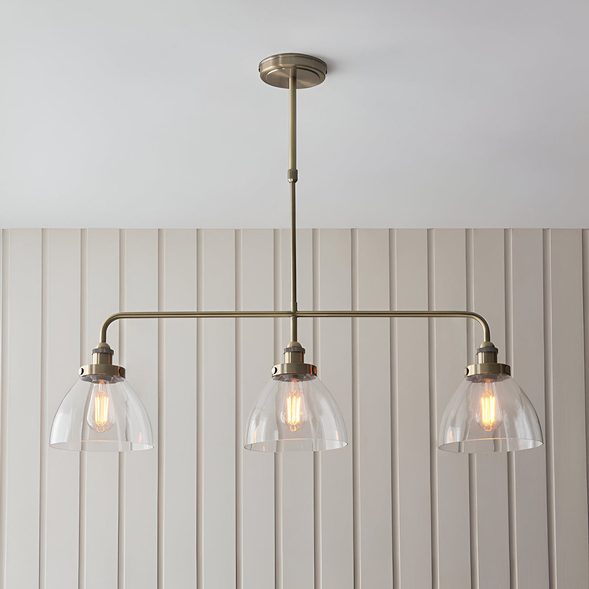 Endon Wentworth 3 light linear stem pendant in antique brass with three clear glass dome shades and exposed filament bulbs, mounted below a panelled wall.