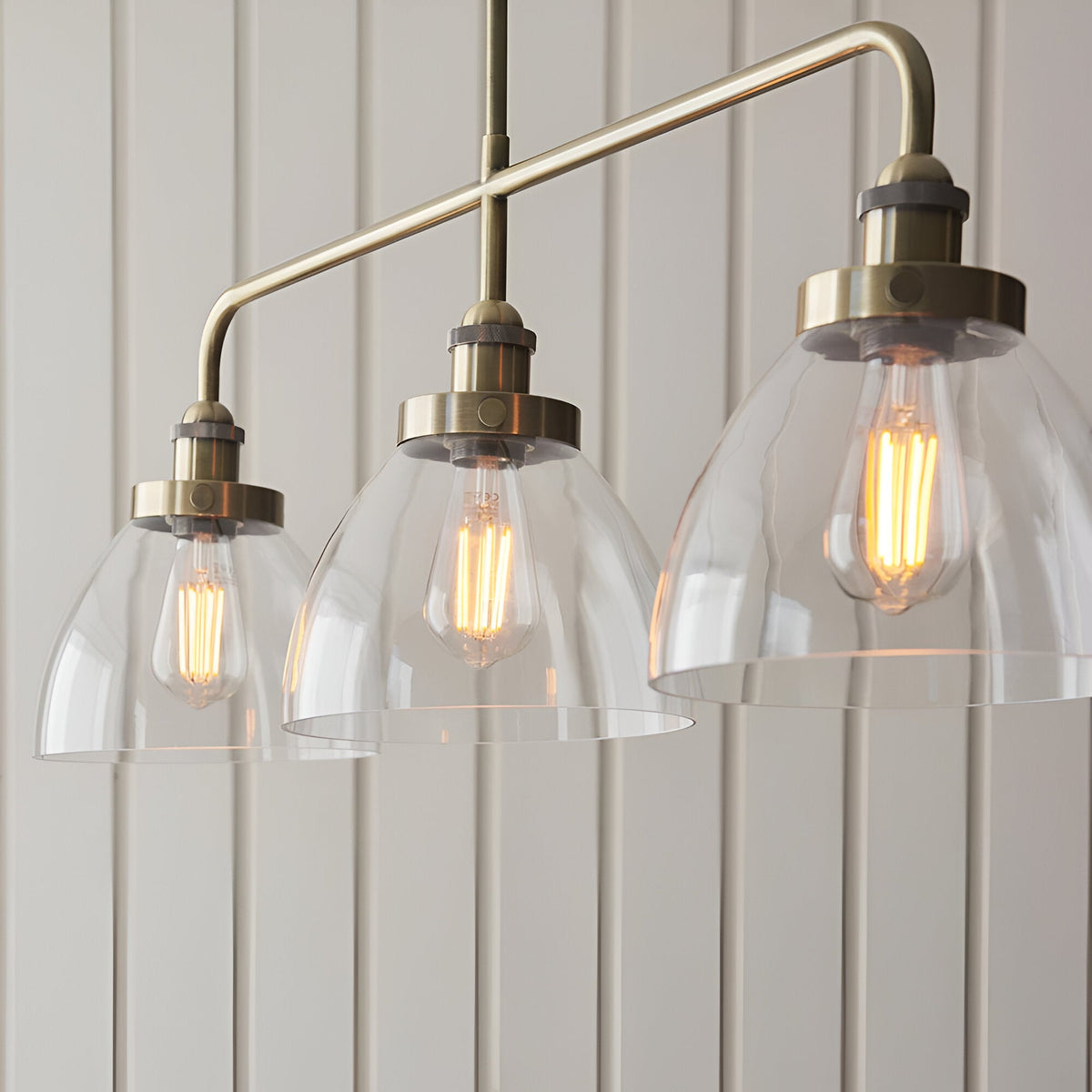 Endon Wentworth 3 light linear stem pendant in antique brass with clear glass dome shades and exposed filament E27 lamps, shown against panelled walls.