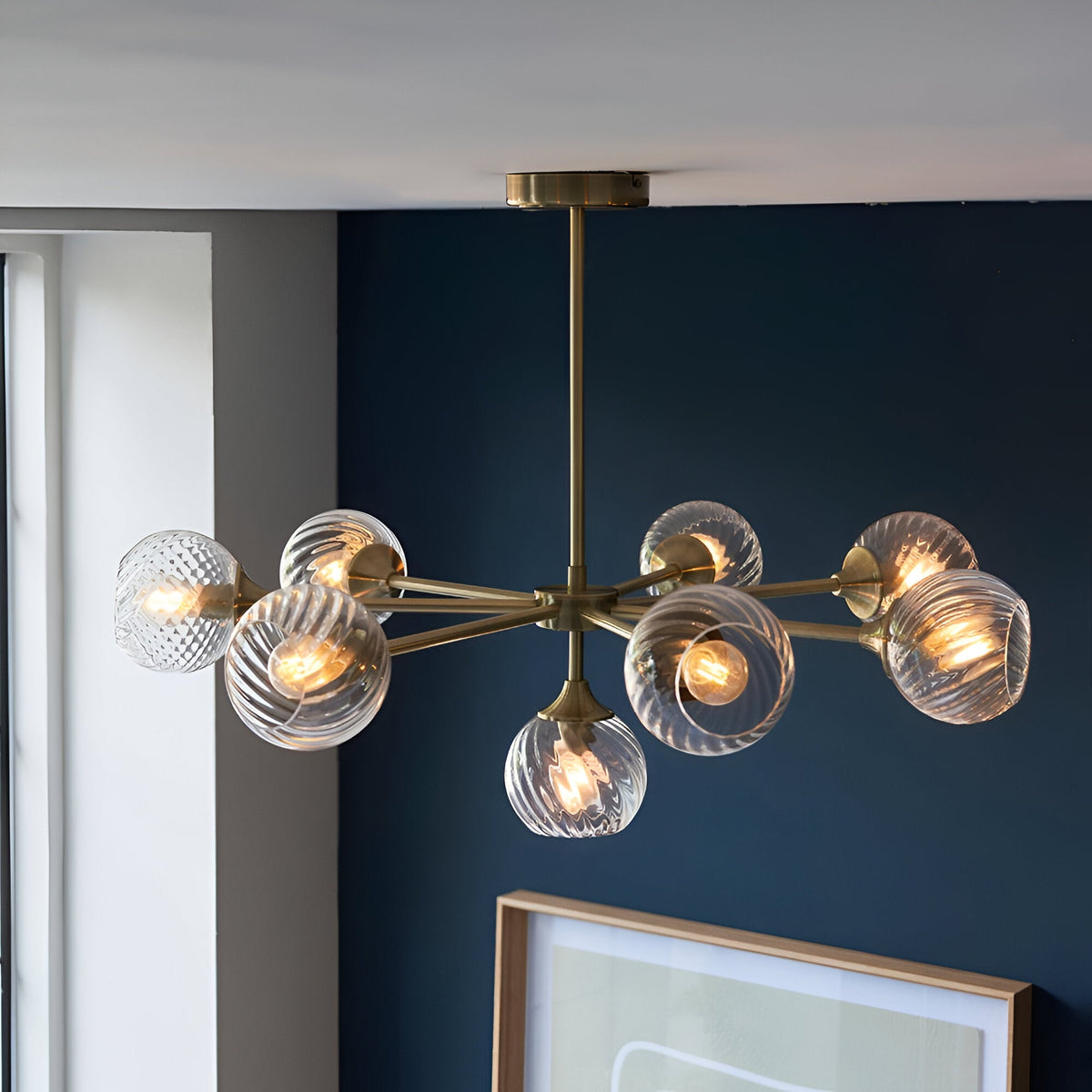 Endon Georgiana 8-light semi-flush pendant in brass with spiral-cut clear glass globe shades, installed against a deep blue wall above a framed print.
