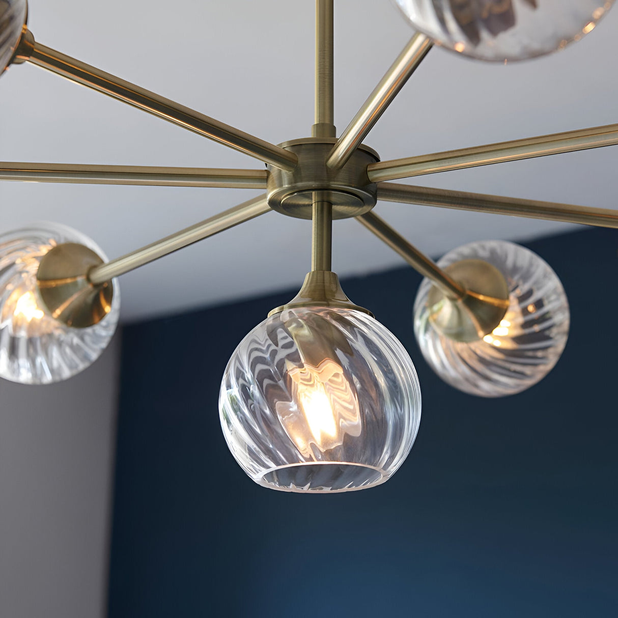 Endon Georgiana 8 light pendant shown close-up with brass spoke arms and spiral-cut clear glass globe shades against a navy and grey wall.