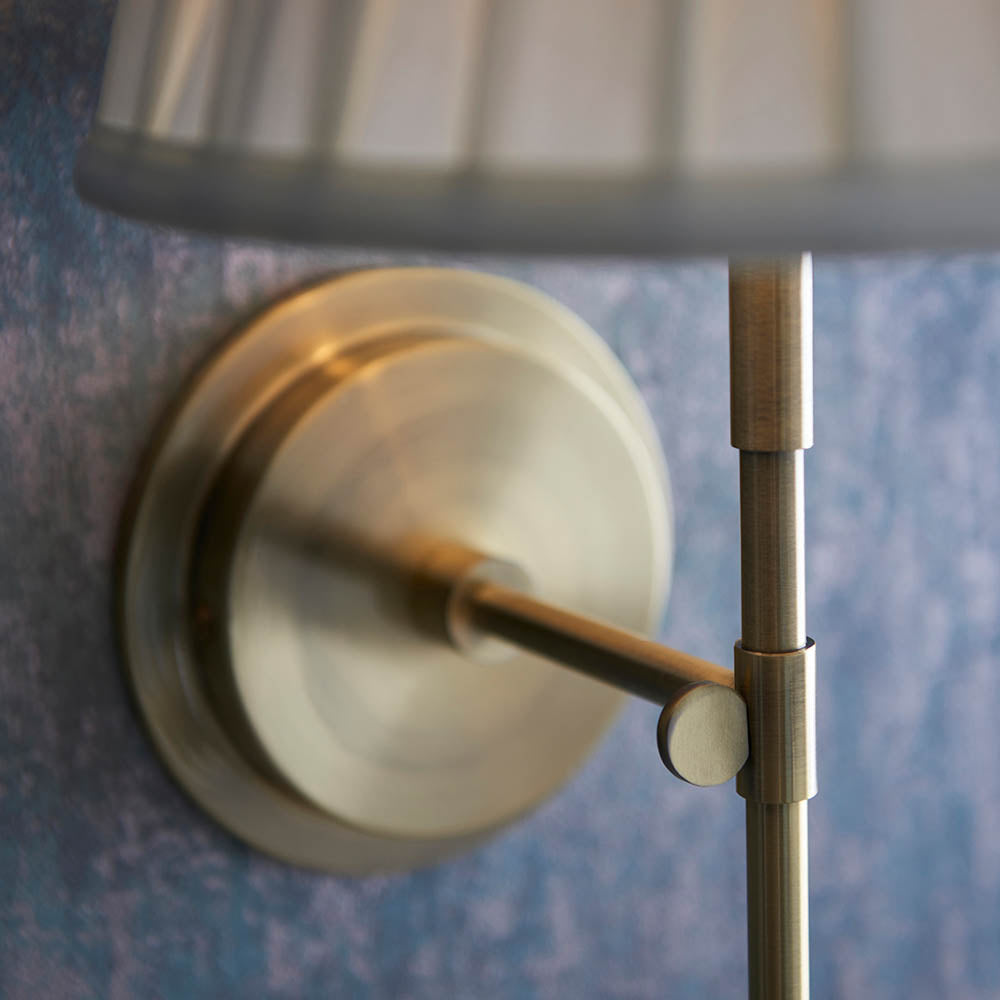 Close-up of the Endon Chartwell wall light's antique brass bracket and adjustable joint against a mottled blue wall, with the pleated cream shade above.