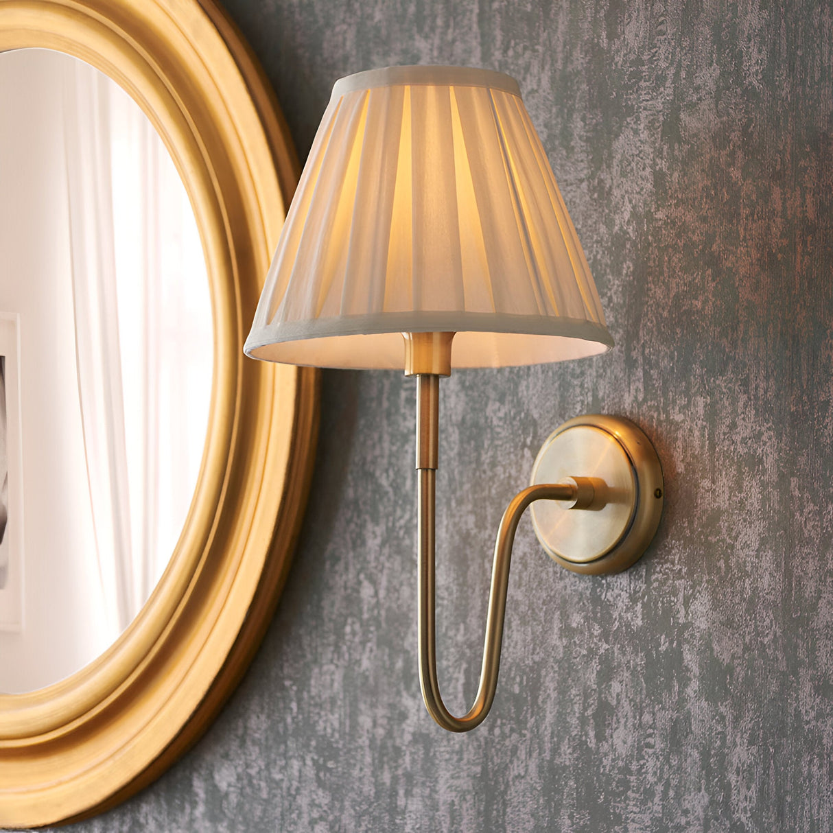 Endon Cliffdon wall light in antique brass with cream box-pleat tapered shade, mounted beside a round gold-framed mirror on a textured grey wall.