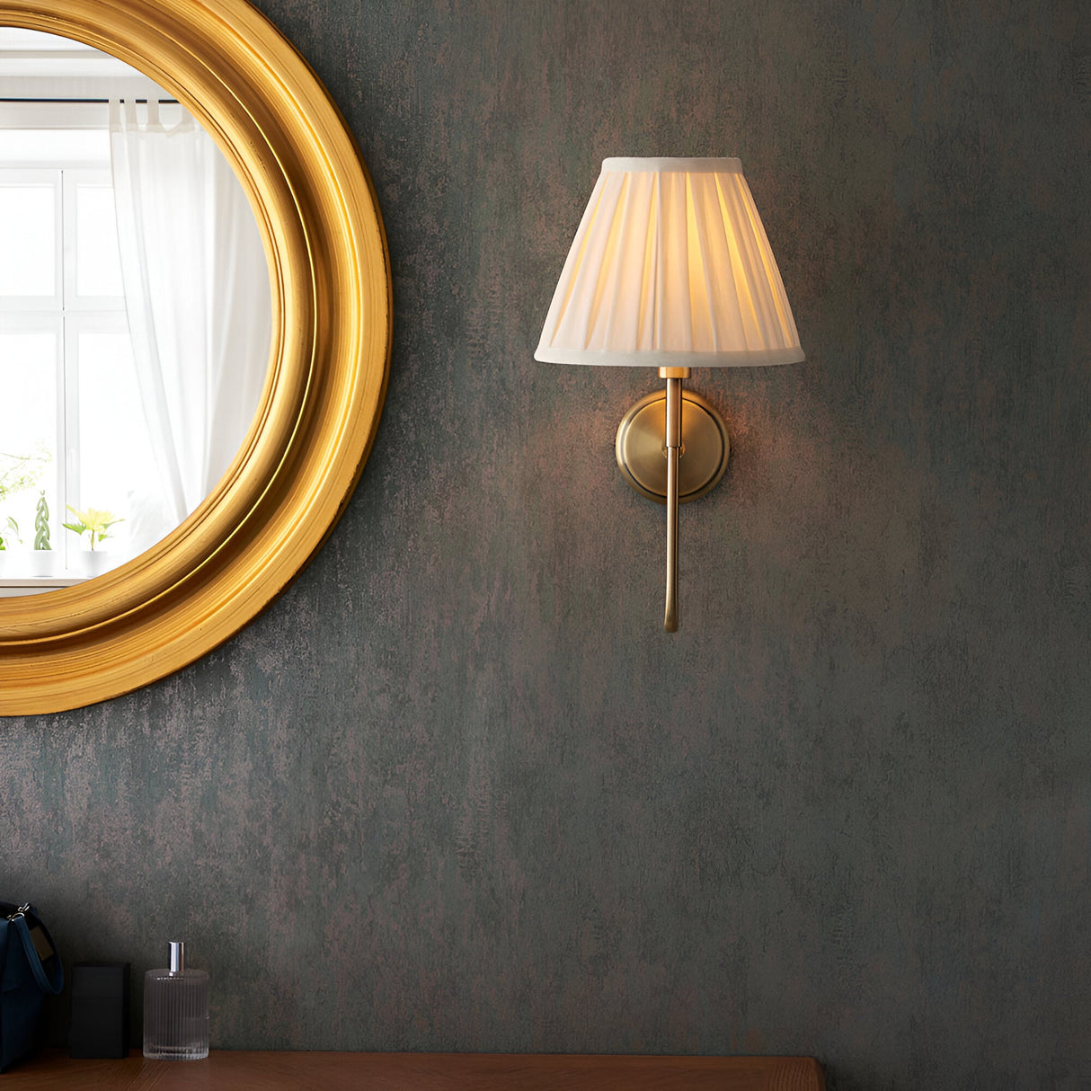 Endon Cliffdon wall light in antique brass with tapered cream box-pleat shade, mounted beside a round gold-framed mirror on a dark textured wall.