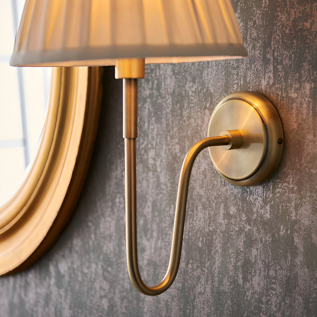 Close-up of the Endon Cliffdon antique brass wall light showing the curved arm, round backplate and pleated cream shade beside a gold-framed mirror.