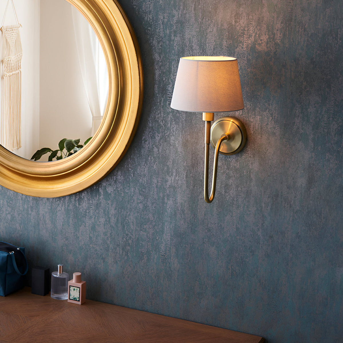 Endon Cliffdon antique brass wall light with tapered ivory fabric shade, mounted on a textured blue wall beside a round gold-framed mirror.