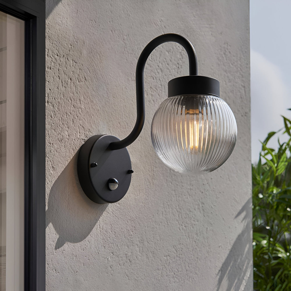 Endon Brackgrove outdoor wall light in black with arched arm, ribbed clear glass globe shade and PIR sensor, mounted on a rendered exterior wall.