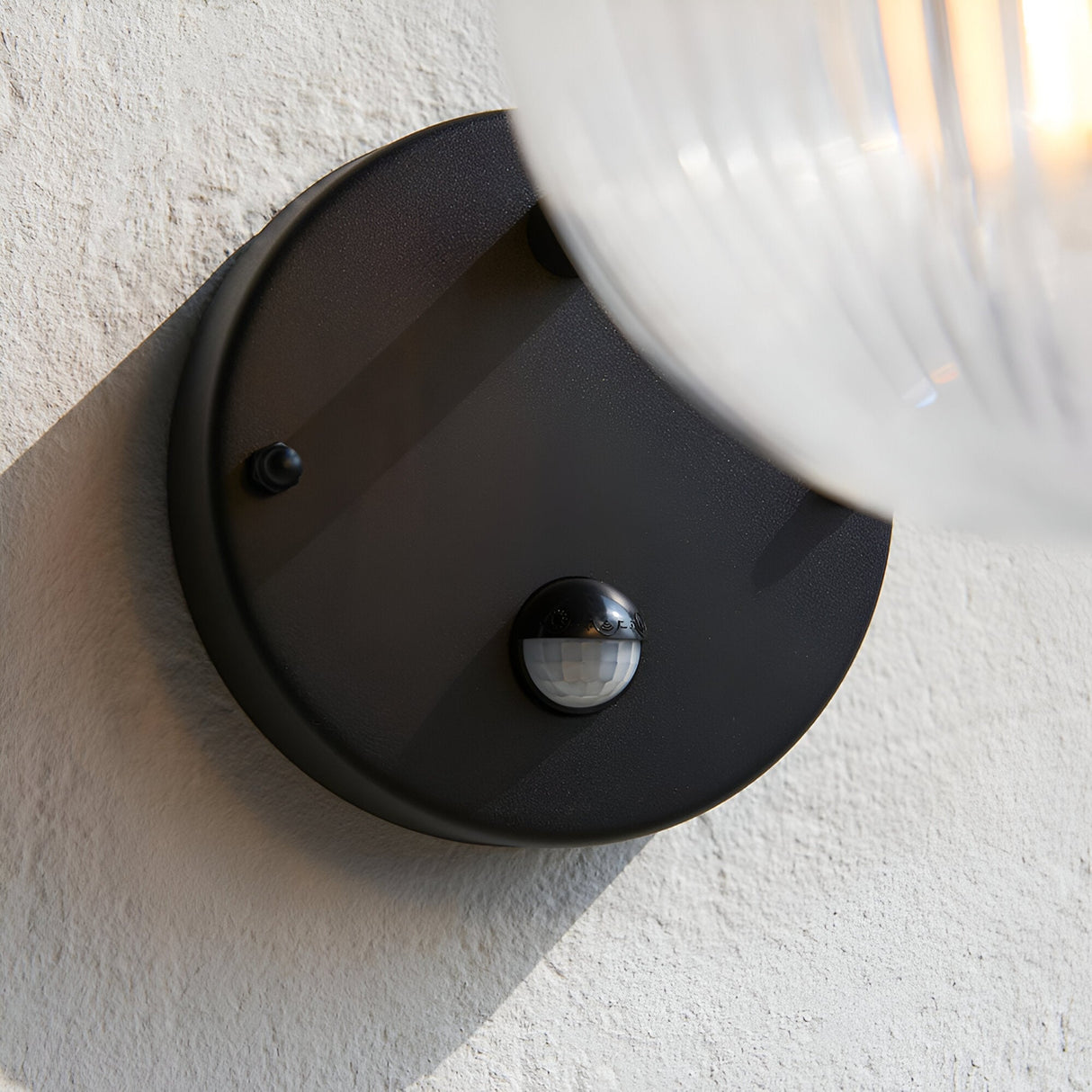 Endon Brackgrove outdoor wall light with textured black round backplate, PIR sensor and clear ribbed glass shade, mounted on a white textured wall.