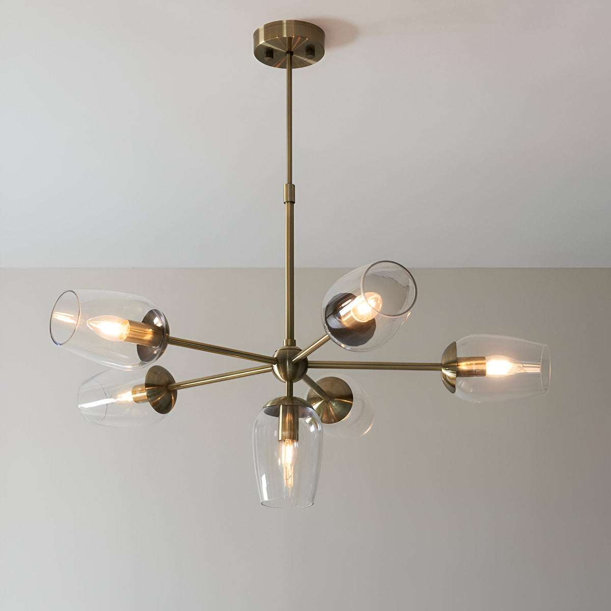 Caldworth 6 Light Pendant in brass with slender curved arms and clear glass shades, hanging from a telescopic stem against a plain light wall.