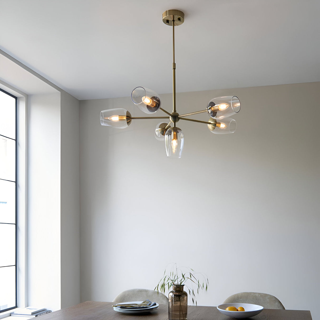 Caldworth 6 Light Pendant in brass with six curved arms and clear glass shades, hanging above a dining table in a neutral-toned room.