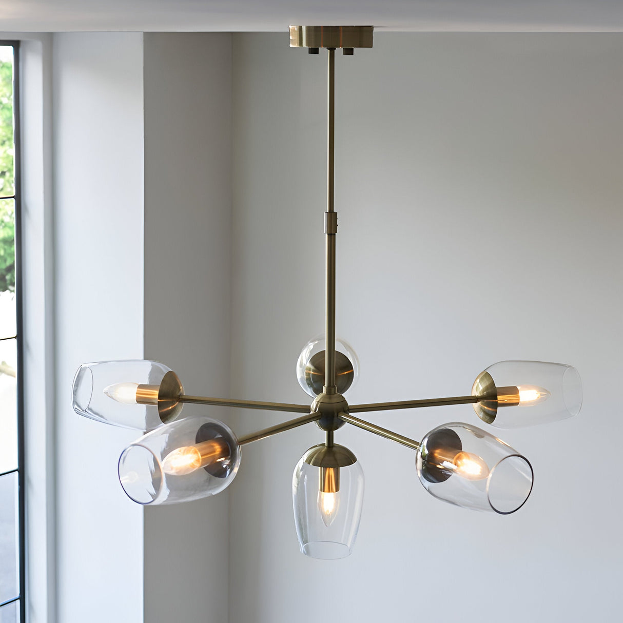 Caldworth 6 Light Pendant in brass, six curved arms with clear glass shades and exposed bulbs, hung from a telescopic stem in a light-coloured room.