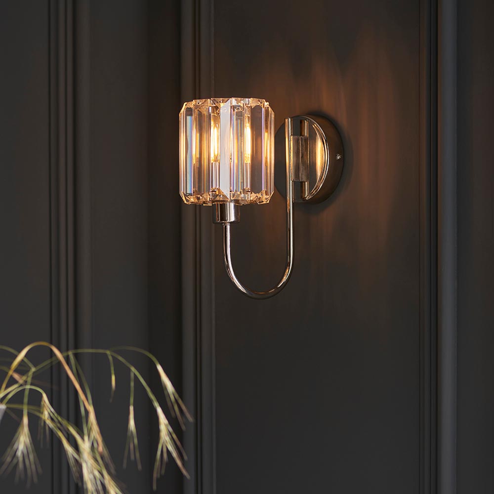 Endon Clementine wall light in nickel with a faceted cut glass shade on a curved plated arm, illuminated against a dark panelled wall.