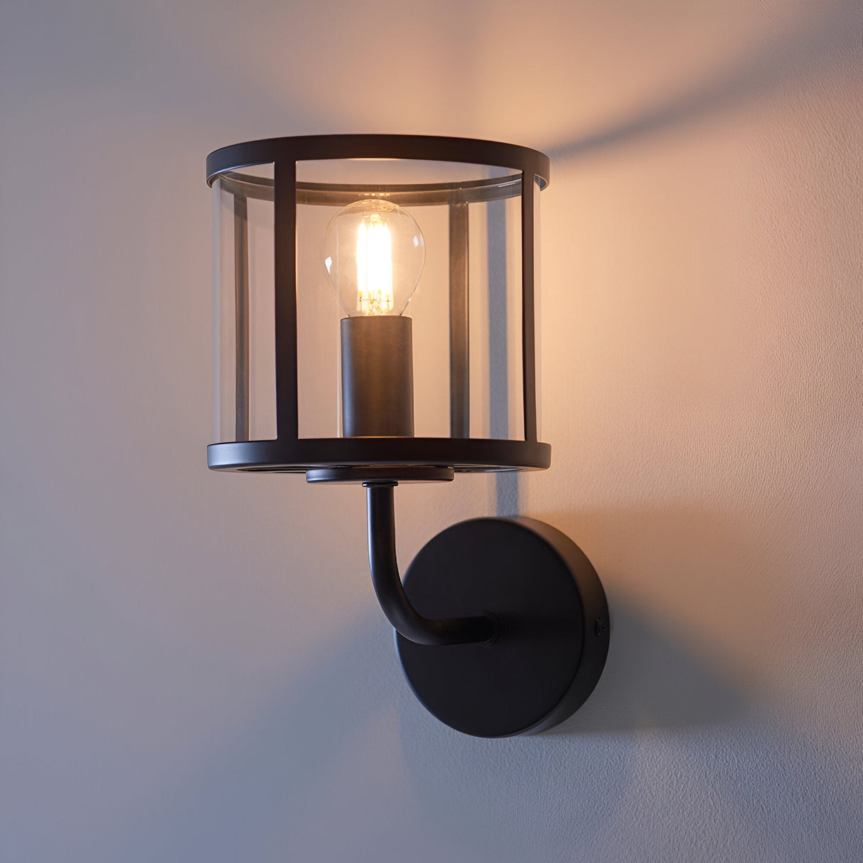 Endon Ellery wall light in matt black with a clear cylindrical glass shade and single E14 filament bulb, mounted on a pale interior wall.