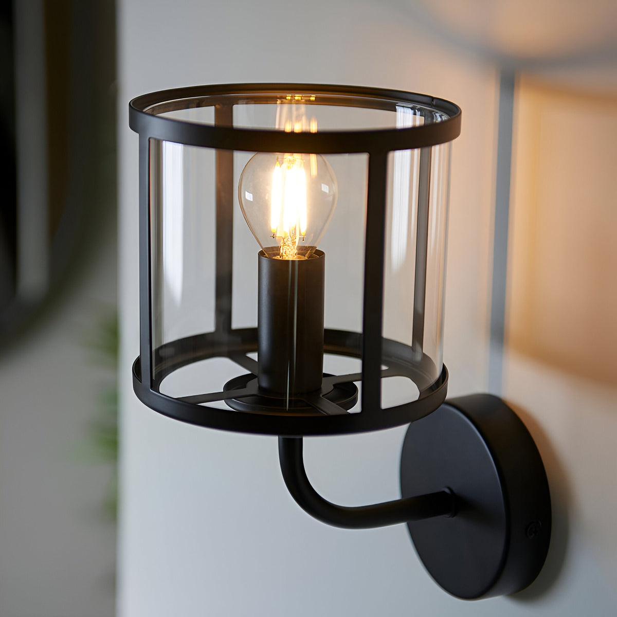 Endon Ellery wall light in matt black with a clear cylindrical glass shade and single E14 filament bulb, mounted on a pale interior wall.