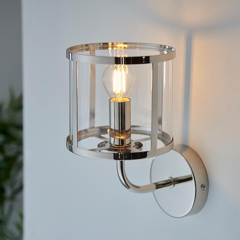 Ellery Wall Light in nickel with a cylindrical clear glass shade and exposed bulb, mounted on a white wall via a curved arm and round backplate.
